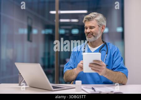 Reifer männlicher Arzt in blauen Peelings und Stethoskop lächelnd, während er ein Tablet hält, und bietet fachkundige Gesundheitsversorgung durch moderne Telemedizin-Technologie von seinem Büro aus Stockfoto