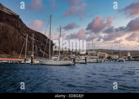 Segelboote und Yachten ruhen in sanftem Abendlicht an schwimmenden Docks aus, während sich die rosa Wolken über vulkanische Klippen, einem Leuchtturm und einer Terrasse ziehen Stockfoto