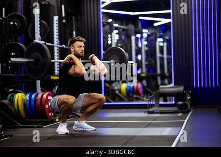 In einem modernen Fitnessstudio mit bunten Gewichten führt ein Mann eine Langhantelhocke aus. Er konzentriert sich intensiv und zeigt Stärke und richtige Form unter bri Stockfoto