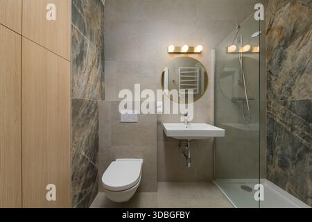 Moderne Inneneinrichtung des Badezimmers mit beigefarbenen und dunklen Steinfliesen. Der Raum umfasst eine begehbare Glasdusche, eine wandmontierte Toilette, ein Rechteck Stockfoto