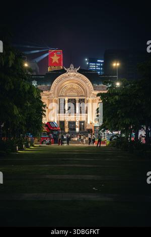 Nächtlicher Blick auf das beleuchtete Saigon Opernhaus in Ho Chi Minh City mit rotem Touristenbus und Wanderern. Moderner Wolkenkratzer im Hintergrund. Stockfoto
