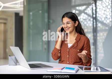 Glückliche junge indische Geschäftsfrau lächelt und spricht mit einem Mobiltelefon, während sie mit einem Laptop und einem Planer an ihrem Schreibtisch sitzt und aktiv kommuniziert Stockfoto