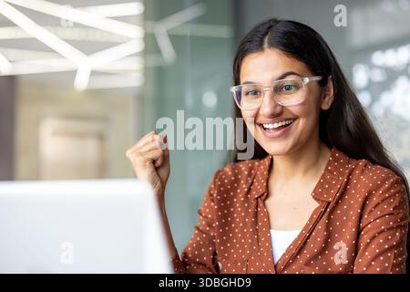 Junge indianerin, die eine Brille trägt und begeistert ist und Erfolg feiert, während sie auf ihren Laptop-Bildschirm blickt, von zu Hause aus oder in einem modernen Büro arbeitet Stockfoto