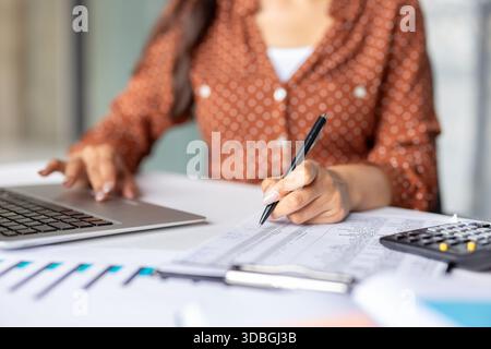 Frau, die Geschäftsfinanzen verwaltet, Ausgaben berechnet und Daten auf einem Laptop überprüft, während sie mit einem Stift und Taschenrechner auf einem Schreibtisch Unterlagen analysiert Stockfoto