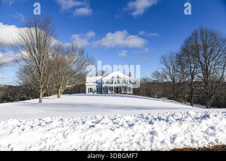 Ein großes weißes Haus mit einem Kamin sitzt auf einem verschneiten Feld. Das Haus ist von Bäumen umgeben und der Himmel ist blau Stockfoto