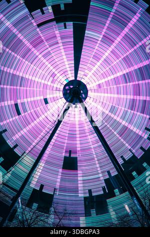 Langzeitaufnahme eines beweglichen Riesenrads auf dem Glasgow Winter Market, das aus dem Foto mit langsamer Verschlusszeit leuchtende kreisförmige Lichtspuren in Pink und Petrol erzeugt Stockfoto