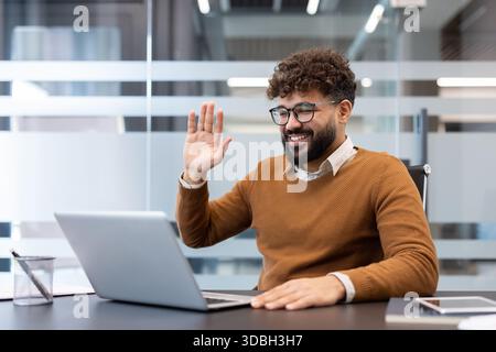 Ein junger Geschäftsmann, der an einem virtuellen Meeting teilnimmt, lächelt und seine Kollegen mit einem freundlichen Wink von seinem modernen Bürotisch aus begrüßt, der sich für Fernkommunikation einsetzt Stockfoto