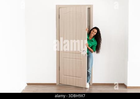 Fröhliche junge Frau, die Besucher an ihrem modernen Apartment-Eingang begrüßt Stockfoto