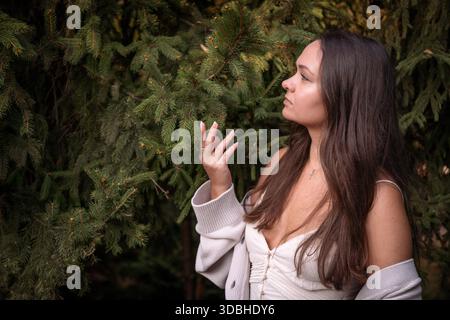 Junge Frau steht neben immergrünen Ästen, sanft berührende Kiefernnadeln, sanftes Tageslicht, tiefe Grüntöne, besinnliche Stimmung Stockfoto