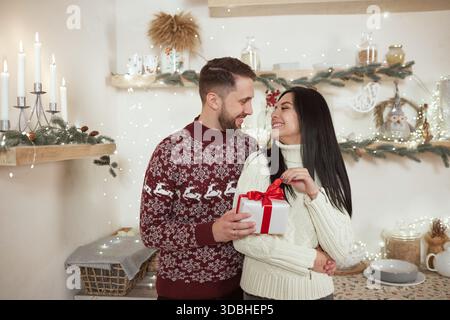 Ein lächelnder Mann schenkt glücklicher Frau in einem gemütlichen, eingerichteten Haus während des Winterferienabends ein gewickeltes Geschenk Stockfoto