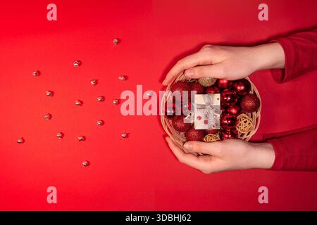 Festliche Komposition mit einem Korb gefüllt mit roten und goldenen Weihnachtskugeln und einer kleinen Geschenkbox, auf rotem Hintergrund mit silbernem Bell angeordnet Stockfoto