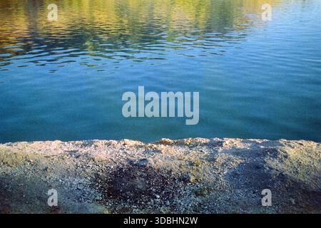 Blick auf den See mit gelbem Uferboden im Vordergrund und blauem Wasser, gegenüber dem Ufer reflektiert, aufgenommen auf analogem 35-mm-Film. Stockfoto