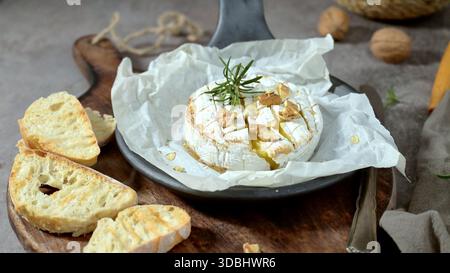 Eine rustikale Nahaufnahme mit geschmolzenem Camembertkäse mit Honig, Walnüssen und frischem Rosmarin. Das Gericht wird auf Pergamentpapier mit knusprig gerösteten Gerichten serviert Stockfoto