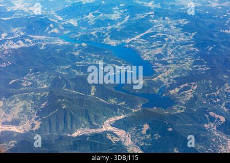 Aus der Vogelperspektive zeigt ein sich windendes blaues Reservoir, das durch bewaldete Hügel und Täler schneidet. Flickenteppich und entfernte Siedlungen schaffen ein riesiges Gelände Stockfoto