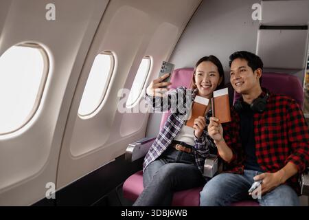 Ein junges asiatisches Paar, das im Flugzeug sitzt und lächelt, während er Pässe und ein Smartphone hält. Sie scheinen aufgeregt und bereit für ihre Reise zu sein, kleidet sich Stockfoto