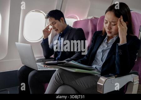 Zwei gestresste asiatische Geschäftsleute sitzen in einem Flugzeug und halten den Kopf frustriert. Der Mann schaut auf den Laptop-Bildschirm, während die Frau Stockfoto
