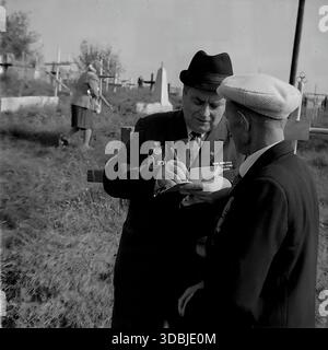 September 1978. Dorf Dolina, Bezirk Sloviansk, Region Donezk, ukrainische SSR, UdSSR. Zwei ältere Männer, wahrscheinlich Veteranen des Zweiten Weltkriegs, unterhalten sich auf einem örtlichen Friedhof während des 35. Jahrestages der Befreiung des Dorfes von der Nazi-Besatzung. Ein Mann, der einen Fedorahut und viele Dienstmedaillen auf seiner dunklen Anzugsjacke trägt, schreibt Kontaktdaten in ein kleines Notizbuch, während der andere ansieht. Im Hintergrund spaziert eine Frau zwischen den hölzernen Grabkreuzen. Authentisches Archivbild, das die Atmosphäre der Erinnerung, der Bruderschaft und des friedlichen Lebens im sowjetischen Donbass erfasst. Stockfoto