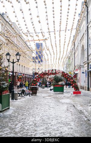 Moskau, Russland, 24.10.2025 festliche Straße dekoriert mit Weihnachtslichtern und rotem Girlandenbogen an einem verschneiten Wintertag mit Menschen, die für den Urlaub gehen c Stockfoto