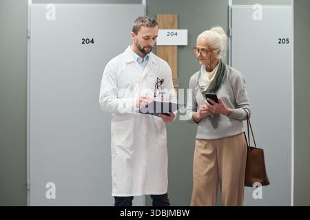 Kaukasischer männlicher Arzt, der medizinische Informationen mit einer älteren kaukasischen Frau bespricht, die Smartphone und Handtasche in der Hand hält und beide im Krankenhausflur in der Nähe nummerierter Türen stehen Stockfoto