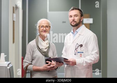 Ältere kaukasische Frau, die neben dem jungen kaukasischen männlichen Arzt steht, der ein Klemmbrett hält, beide auf die Kamera im Flur der medizinischen Einrichtung schauen und sich beraten lassen Stockfoto