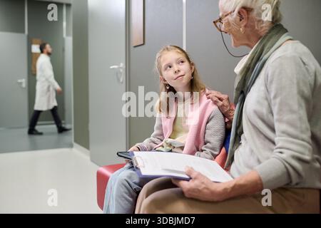 Kaukasische ältere Frau, die mit dem kaukasischen Mädchen Kind spricht, während sie im medizinischen Wartebereich sitzt, Mädchen aufmerksam zuhört, Doktor im Hintergrund geht, Zwischenablage in Frauenhänden Stockfoto