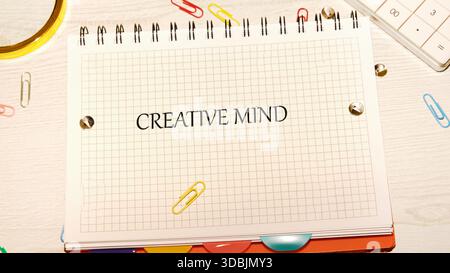 Ein Notizbuch mit Rastermuster liegt offen, auf den Seiten sind die Worte Creative Mind gedruckt. Büroklammern, Klebeband, Taschenrechner und anderes Bürozubehör A Stockfoto
