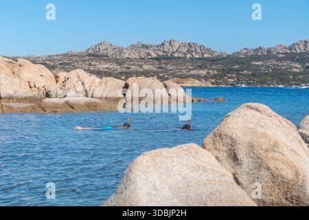 An einem sonnigen Sommertag schwimmen Menschen im Meer nahe einer felsigen Küste. Freizeitaktivität, Urlaub am Meer und natürliche Küstenumgebung, Sardinien Stockfoto