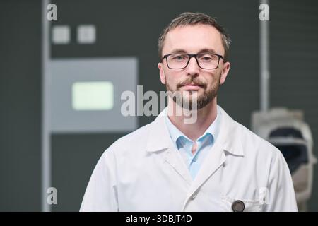 Porträt eines kaukasischen Mannes mittleren Alters mit Brille und Labormantel, der in einer medizinischen Einrichtung steht und direkt in die Kamera blickt, mit neutralem Ausdruck, kurzen Haaren und beschnittenem Bart Stockfoto