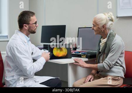 Kaukasischer männlicher Arzt zeigt digitale Netzhautscanner auf Tablette für ältere kaukasische Frau während der medizinischen Beratung, beide sitzen am Schreibtisch im klinischen Büro und besprechen die Augengesundheit Stockfoto