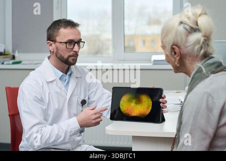 Kaukasischer männlicher Arzt erklärt der älteren kaukasischen Frau während der medizinischen Beratung in der modernen Klinik die Ergebnisse der Netzhautuntersuchung auf einem digitalen Tablet, beide sitzen am Schreibtisch Stockfoto