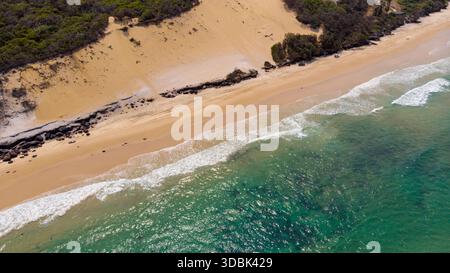 Aus der Vogelperspektive schweifen die schaumigen Wellen über den goldenen Sand des Strandes neben den bepflanzten Sanddünen, Rainbow Beach, Queensland, Australien. Stockfoto