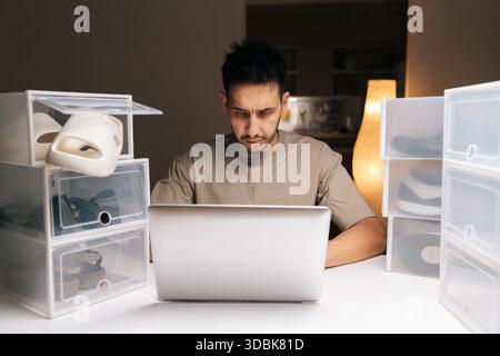 Porträt eines männlichen Unternehmers, der von zu Hause aus einen Online-Schuhladen führt, neue Schuhe in durchsichtigen Aufbewahrungsboxen organisiert und ein Notebook für das Tippen verwendet Stockfoto