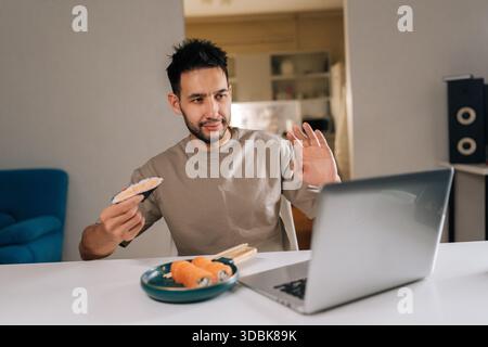 Beschäftigter junger Freelancer, der von zu Hause aus arbeitet, Videoanrufe hat, Onigiri und Sushi-Brötchen in der kurzen Pause isst und Aufgaben mit ausbalanciert Stockfoto