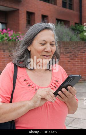 Nahaufnahme einer glücklichen älteren Latina-Frau, die draußen ein Handy benutzt, redet und SMS schreibt. Stockfoto