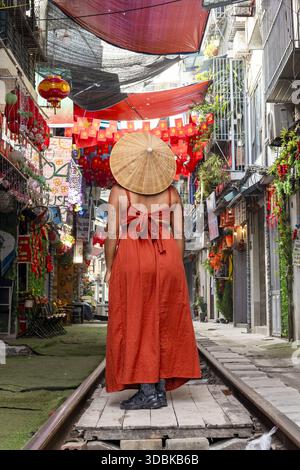 Blick auf eine Frau in orangefarbenem Kleid, die auf Bahngleisen steht, inmitten von leuchtenden roten Laternen und geschäftigem Straßenleben, Hanoi, Hanoi, Vietnam. Stockfoto