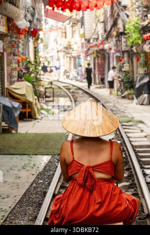 Blick auf eine Frau in rotem Kleid und traditionellem Hut auf Bahngleisen, die durch eine enge Straße mit roten Laternen verlaufen, Hanoi, Hanoi, Vietn Stockfoto