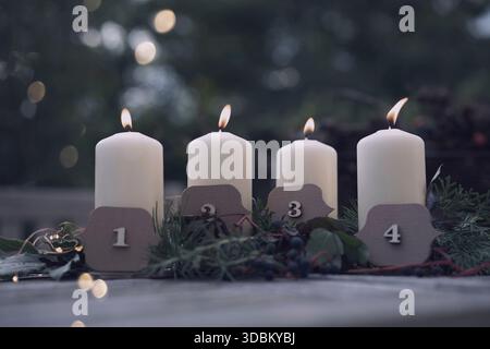 Vier brennende Adventskerzen. Brennende Kerzen für den vierten Advent. weihnachtsdekoration im Freien mit vier weißen Kerzen vor einem Garten und Platz für Text. Stockfoto