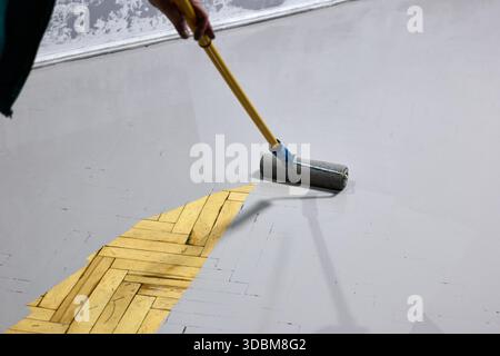Nahaufnahme eines Holzparkettbodens mit grauer Farbe mit einer Rolle, die ein Renovierungsprojekt zeigt. Dieses Bild erfasst den Prozess Stockfoto