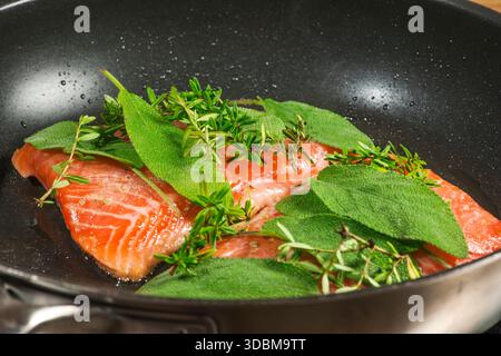 Erhöhte Nahaufnahme zweier Lachsfilets, die mit frischem Salbei und Thymian in einer schwarz beschichteten Pfanne mariniert wurden. Stockfoto