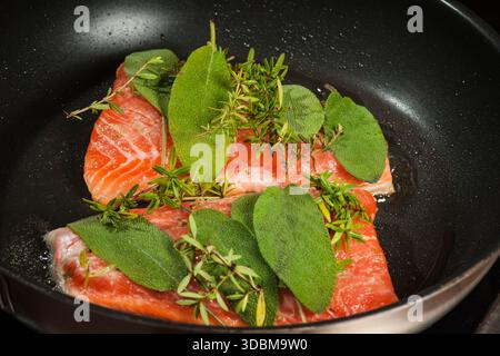 Erhöhte Sicht auf zwei Lachsfilets, die mit frischem Salbei und Thymian in einer schwarz beschichteten Pfanne mariniert wurden. Stockfoto