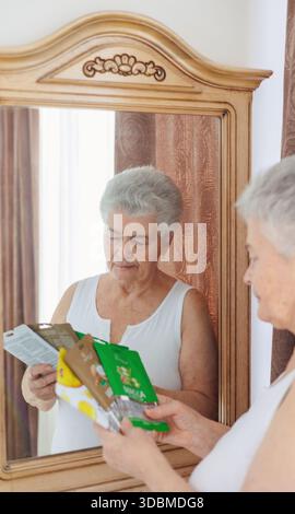 Ältere Frau wählt Gesichtsmaske Kosmetikprodukt, während sie zu Hause vor einem Spiegel steht. Gesunder Lebensstil, achtsames Altern, Selbstpflege, Vitalität Stockfoto