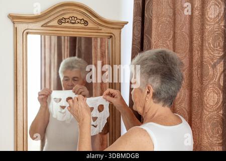 Ältere Frau, die zu Hause Hautpflegemaske vor dem Spiegel aufträgt – Schönheit, Selbstpflege und Anti-Aging-Routine für reife Haut und Wellness-Konzept Stockfoto