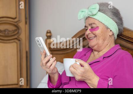 Seniorin 70+ genießt Selbstpflege im Bett mit Augenflecken, Tee und Smartphone, entspannende Morgenroutine. Kopierraum Stockfoto