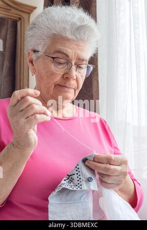 Ältere Frau mit Brille, die am Fenster in einem gemütlichen hellen Schlafzimmer näht, genießt einen ruhigen Moment. Seniorenleben, Sehpflege, handgefertigtes Handwerk Stockfoto