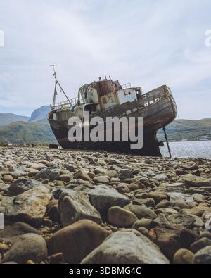 Blick auf das alte, rostige Schiffswrack an einem felsigen Ufer, im Kontrast zum ruhigen blauen Wasser und den fernen majestätischen Bergen, Fort William, Stockfoto