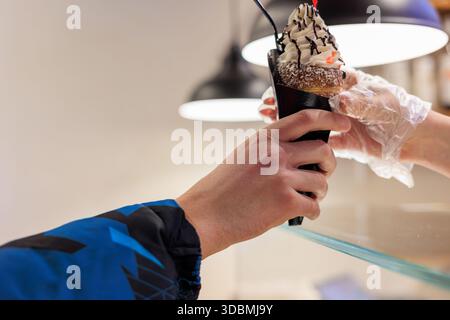 Die Hand hält Trdelnik (Kaminkuchen) in Tasse, Sahne, Schokolade und Kirschgarnitur. Handschuhe des Anbieters sichtbar. Zähler. Beleuchtet den Hintergrund. Street Food Stockfoto