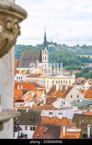 Die Veitskirche steht hoch unter roten Dächern in Cesky Krumlov. Der Sommerhimmel bringt ein sanftes Licht in die historischen Gebäude der Stadt und das üppige Grün im Hintergrund. Stockfoto