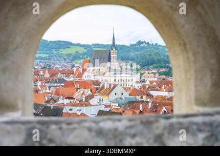 Besucher können die Dächer von Cesky Krumlov durch einen Steinbogen sehen. Die Stadt umfasst Gebäude mit roten Dächern und einem hohen Kirchturm im Hintergrund. Es ist ein Sommertag. Stockfoto