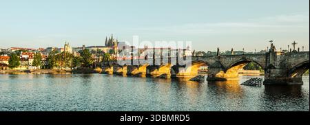 Die Prager Burg erhebt sich majestätisch über der Moldau, während die Karlsbrücke sich anmutig über das Wasser erstreckt. Das Licht am frühen Morgen wirft einen goldenen Farbton über dieses ikonische Stadtbild. Stockfoto