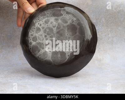 Nahaufnahme einer handgefertigten Keramikplatte mit organischem Glasurmuster, gehalten von einer Hand vor einem neutralen Studiohintergrund. Minimalistisches Stillleben betont Stockfoto